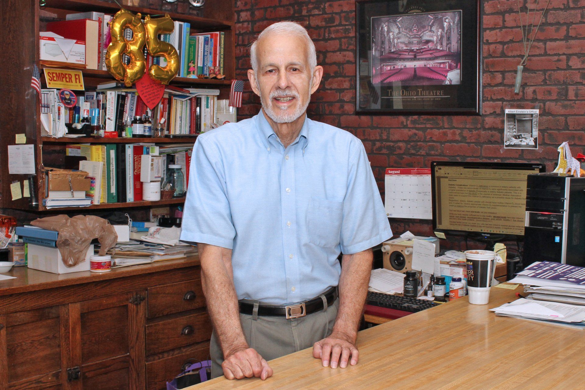An older person in a light blue button-down shirt stands in an office with shelves of books and a computer.