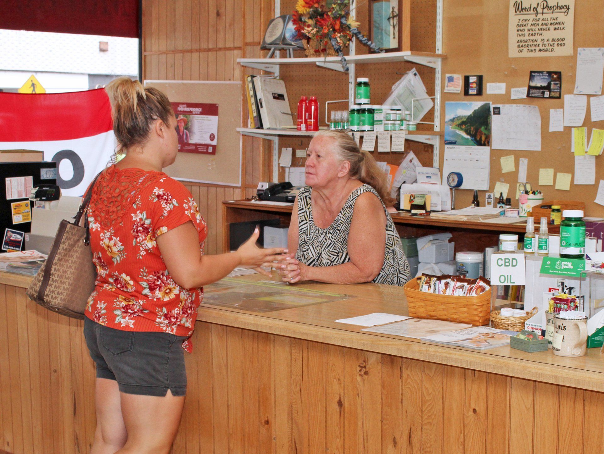 A person in a floral orange top speaks to an individual behind a wooden reception counter in an office setting.