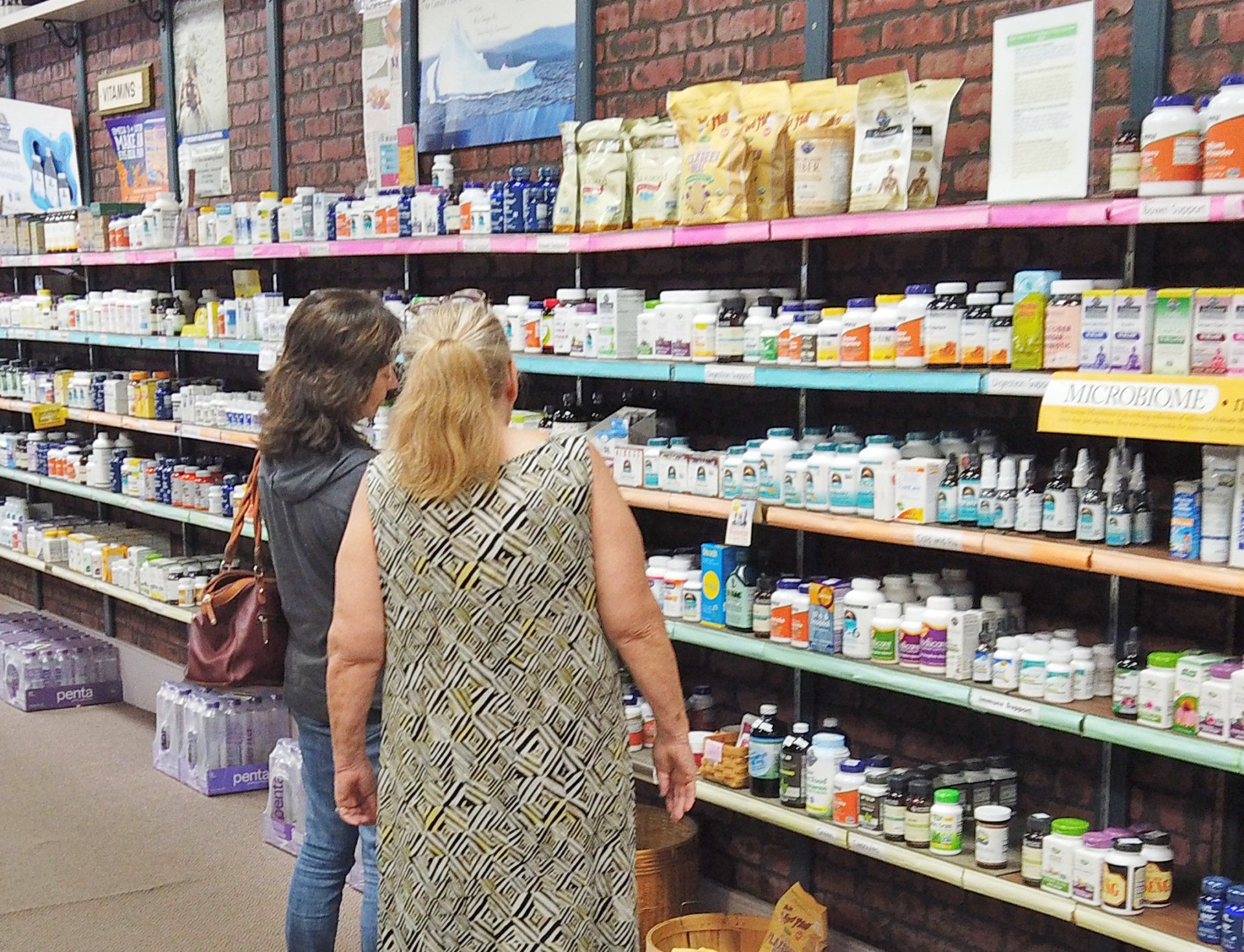 Two people browsing shelves of supplements and vitamins in a store with brick walls.