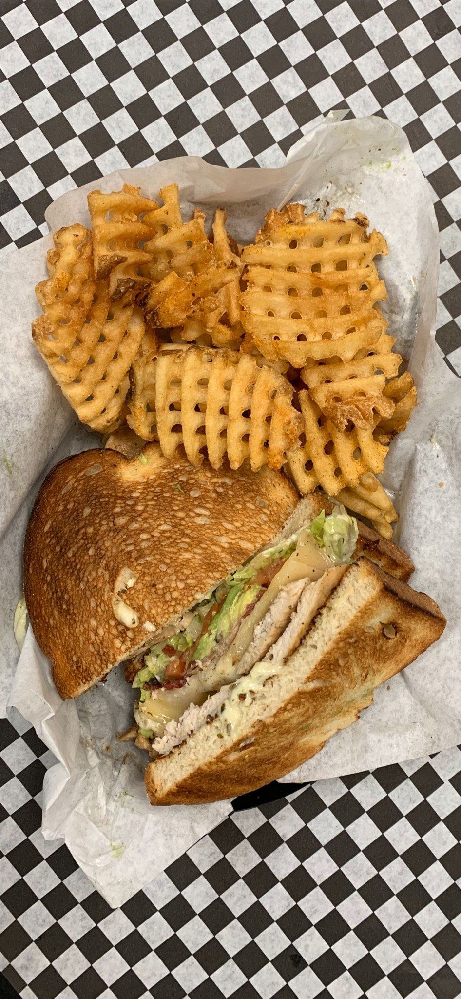 Club sandwich with waffle fries on checkered surface.