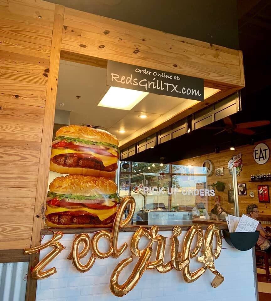 A restaurant, Reds Grill, with burger balloons and a pickup window. Order online at RedsGrillTX.com.