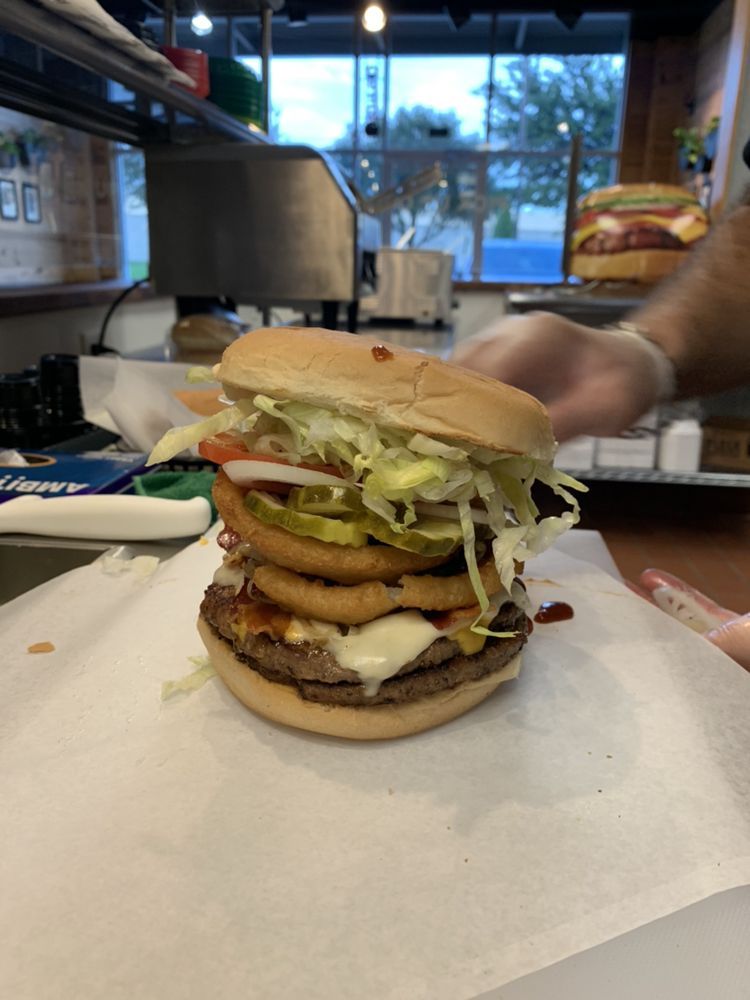 Double-patty burger with cheese, onion rings, pickles, tomato, and lettuce on a bun, being assembled on paper.
