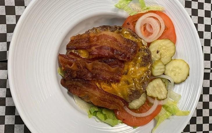 Cheeseburger with bacon and fixings on a white plate; checkered background.