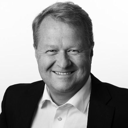 En mann i dress og hvit skjorte smiler på et svart-hvitt bilde.