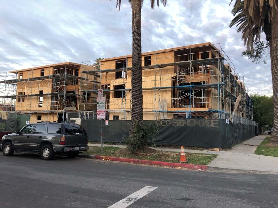 Construction site of multi-story apartment buildings, wooden frames, scaffolding, fenced, parked SUV, palm trees.