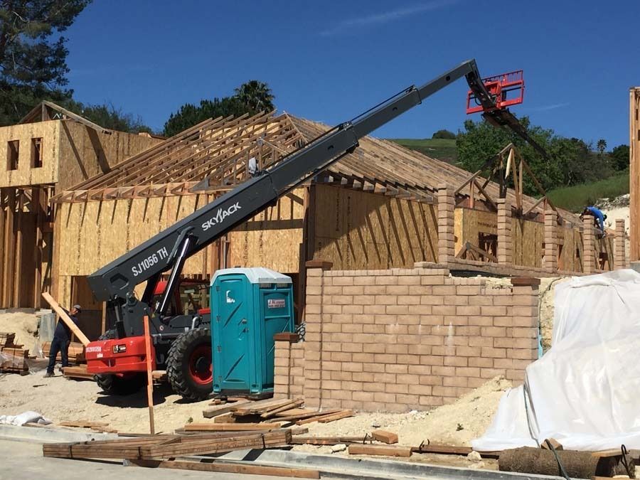 Construction site with a telescopic handler; a person works near a brick wall and wooden structure under a bright blue sky.