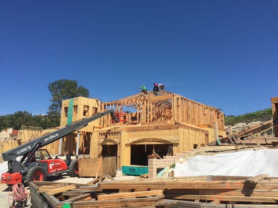 Construction site with workers on a wood-framed house roof. A telehandler lifts materials, clear blue sky.