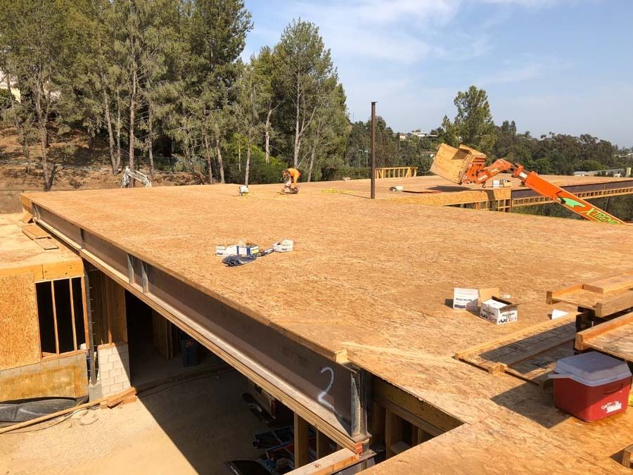 Construction site with plywood platform, steel beams, workers, and surrounding trees.
