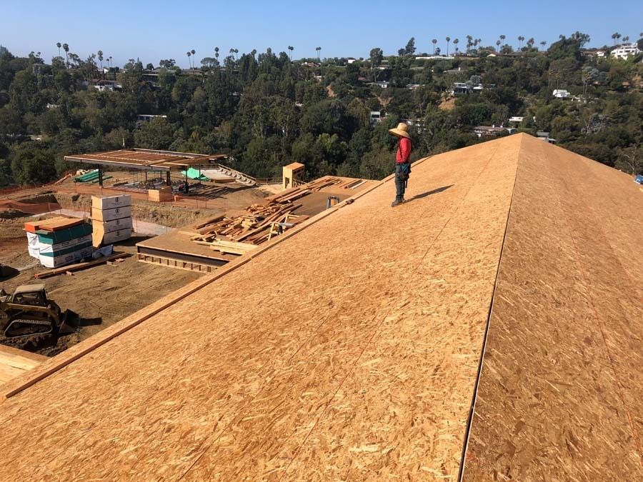 Roofer stands on a wood-covered roof, overlooking a construction site on a hillside with trees under a blue sky.