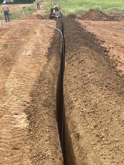 A deep trench cut in brown soil with a black pipe inside; a machine and workers in the background.