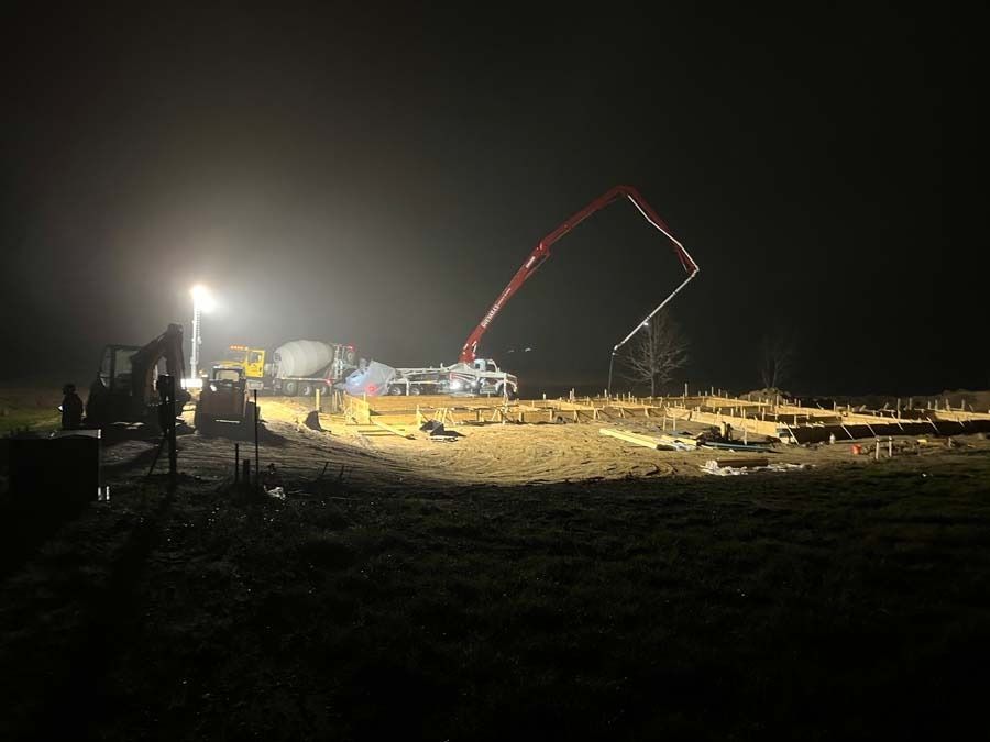 Construction site at night; concrete pump truck pouring concrete onto a foundation, lit by work lights.