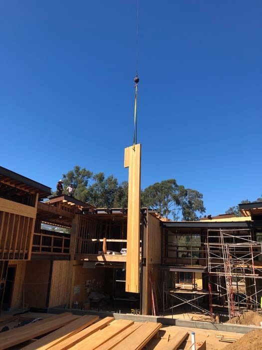 A crane lifting a tall wooden wall panel onto a house under construction on a sunny day.