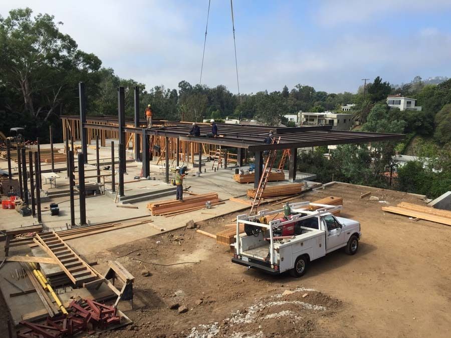 Construction site with steel beams and a white truck. Workers on the structure, surrounded by trees and blue sky.