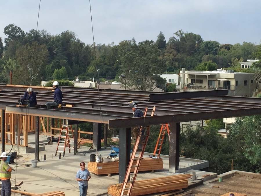 Construction site: workers on a building frame, installing beams. Trees and houses in the background.