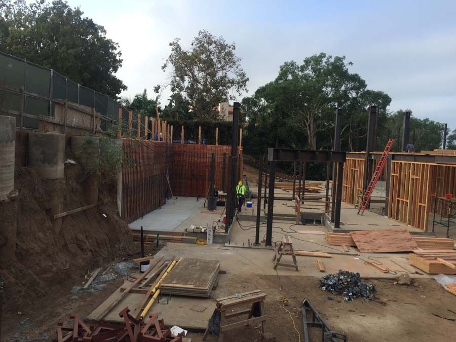 Construction site with concrete foundation, wood framing, and steel supports. A worker in a vest is visible.