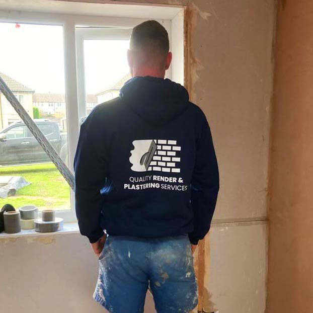 Man in navy hoodie and paint-splattered shorts, standing in a room with exposed plaster.