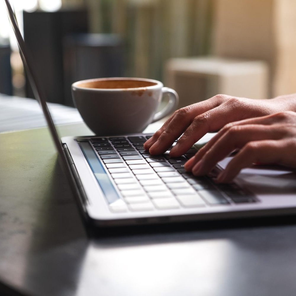 Hands typing on laptop keyboard, coffee cup nearby on table.