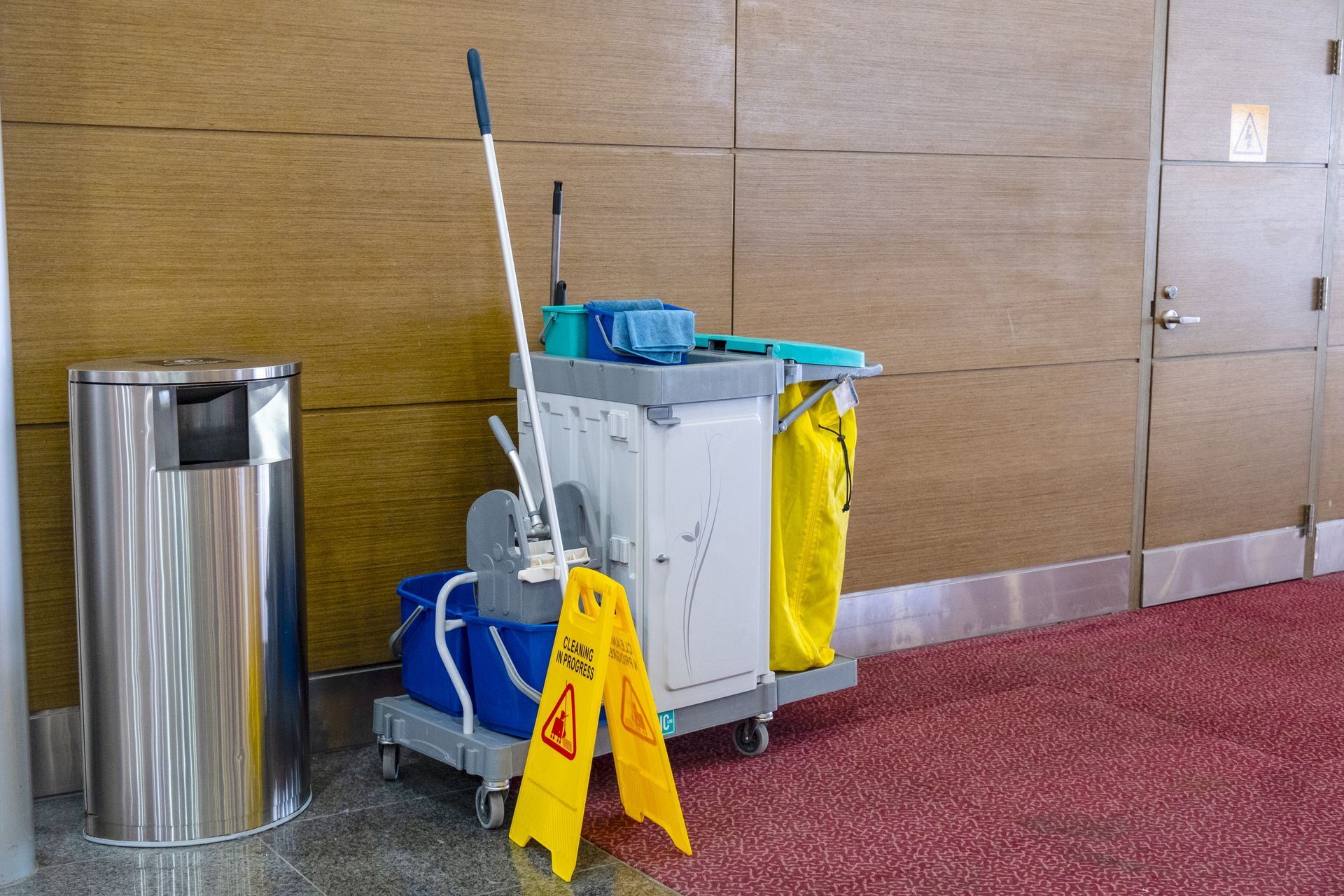 Janitor cart with mop and yellow wet floor sign beside a stainless steel trash can in a hallway