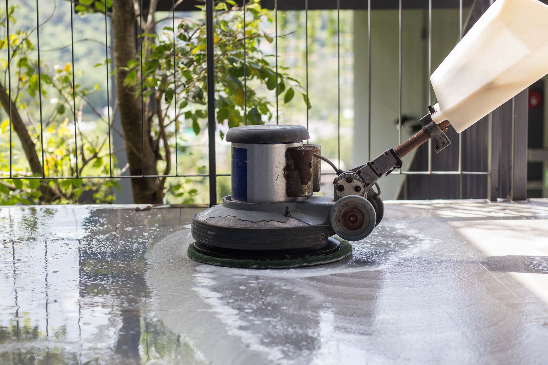 Floor buffer polishing a wet patio surface outdoors near plants and a fence.