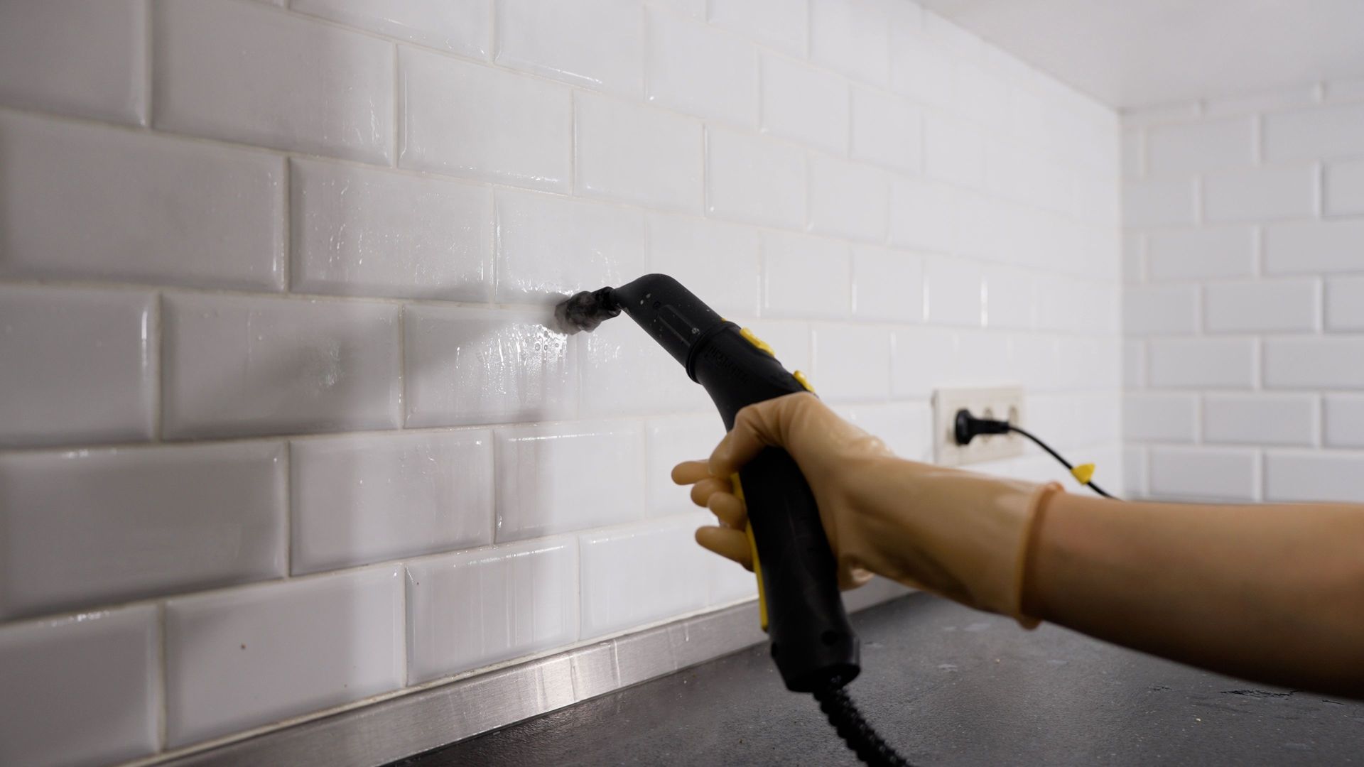 Gloved hand spraying water onto white tiled kitchen backsplash near a countertop and outlet