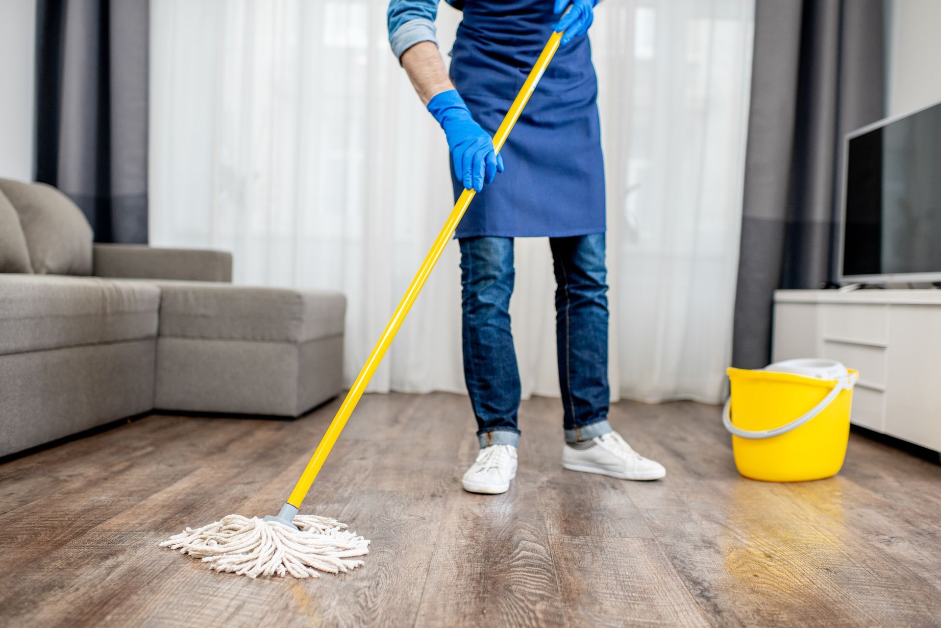 Person mopping a wood floor in a living room beside a yellow bucket