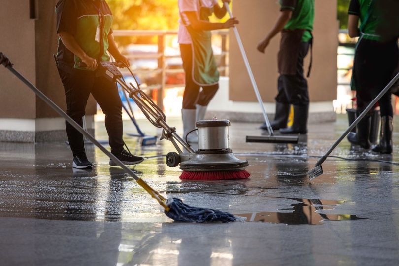 Workers cleaning a wet floor with scrubbers and mops in a sunlit indoor space