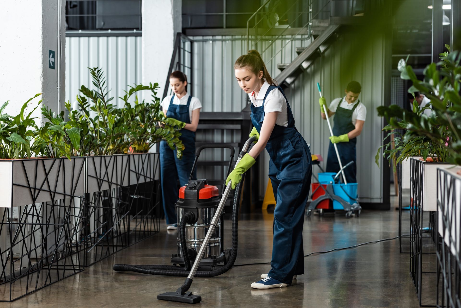 People cleaning a building lobby with vacuum and mop, surrounded by plants and stairs.