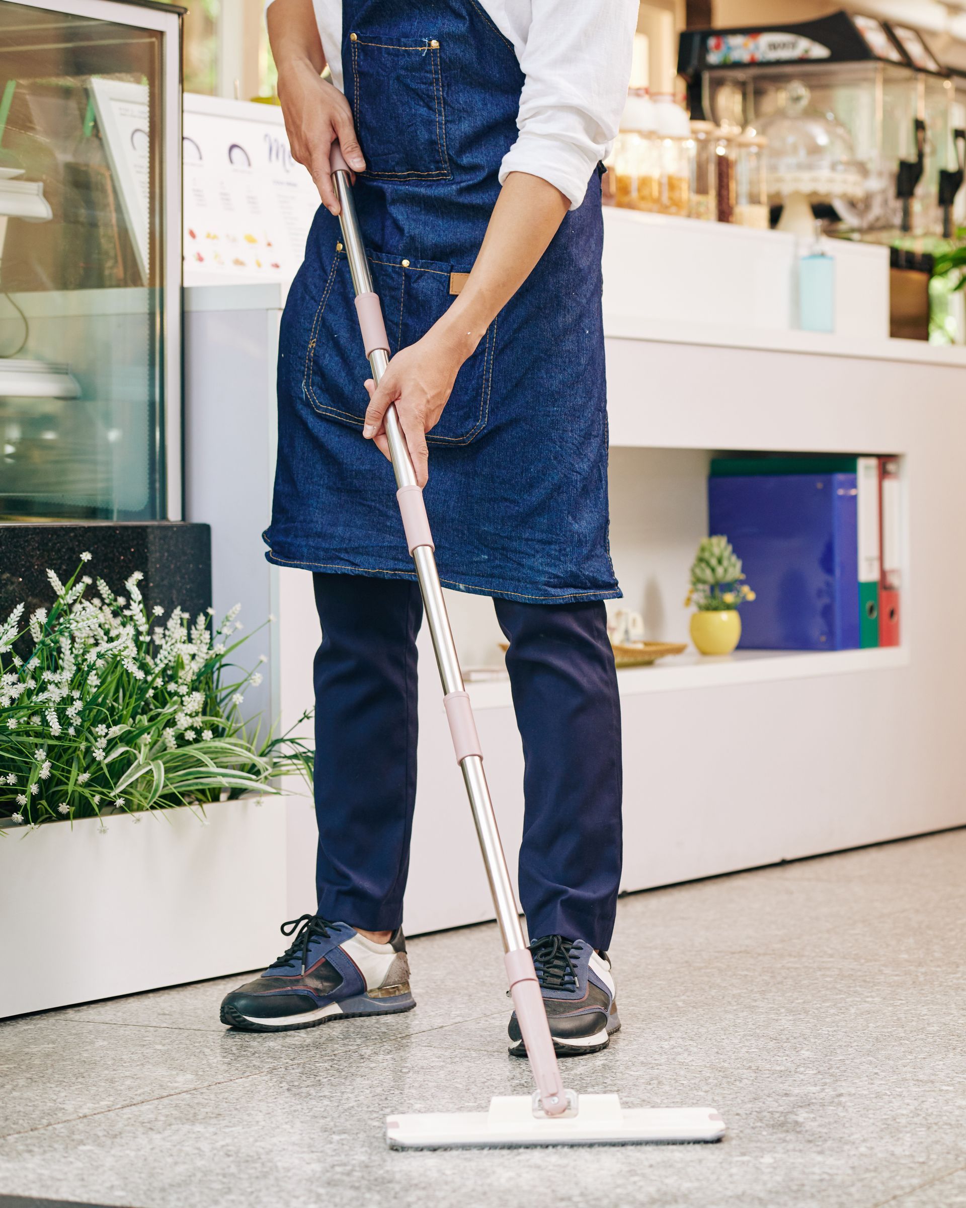 Person mopping a floor in a store aisle with a white mop and denim apron