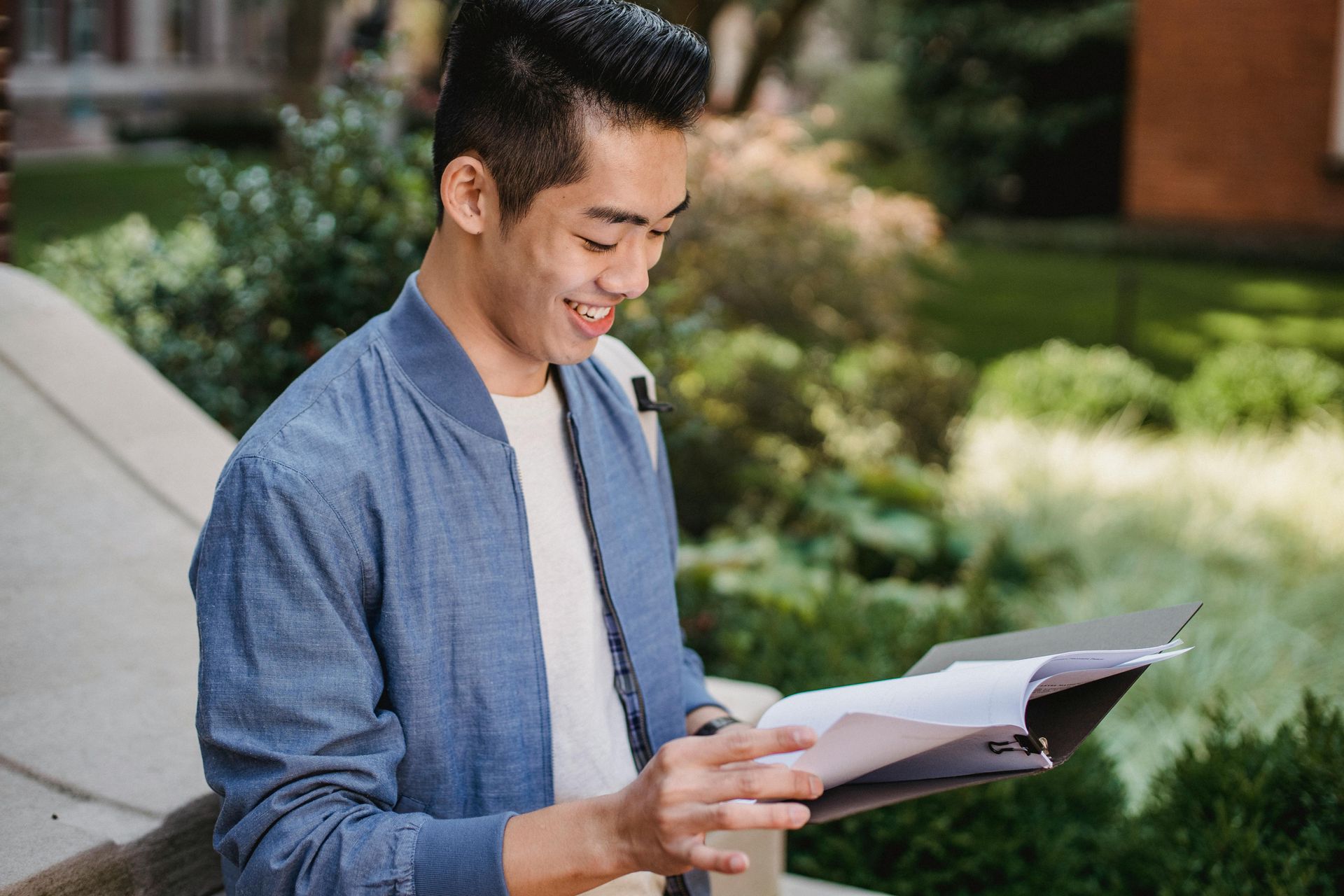 A person wearing a blue bomber jacket over a white shirt smiles while reviewing a document in an outdoor setting.