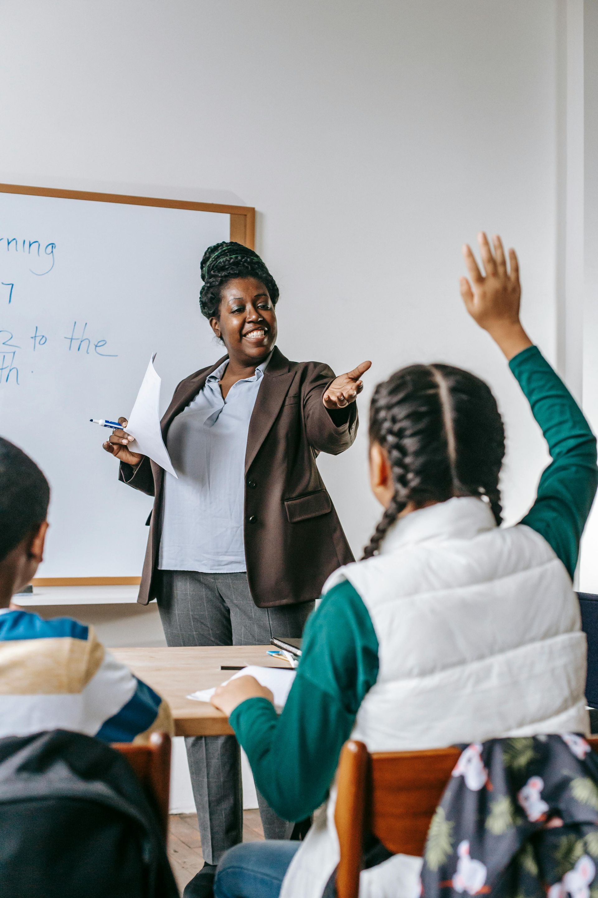 A smiling teacher gestures toward a student with their hand raised in a classroom.