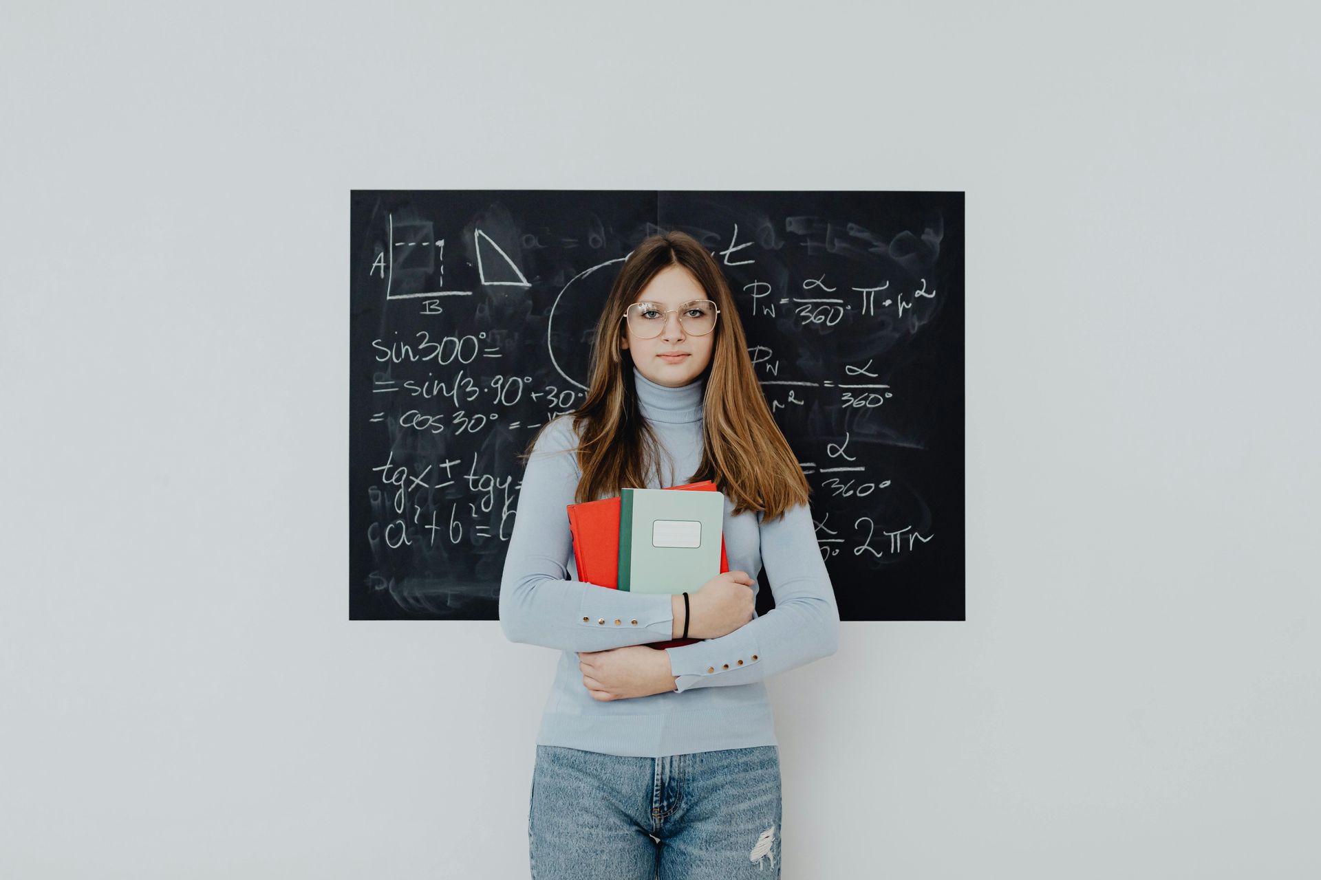 A student in a light blue turtleneck and jeans holds notebooks in front of a chalkboard covered in math formulas.