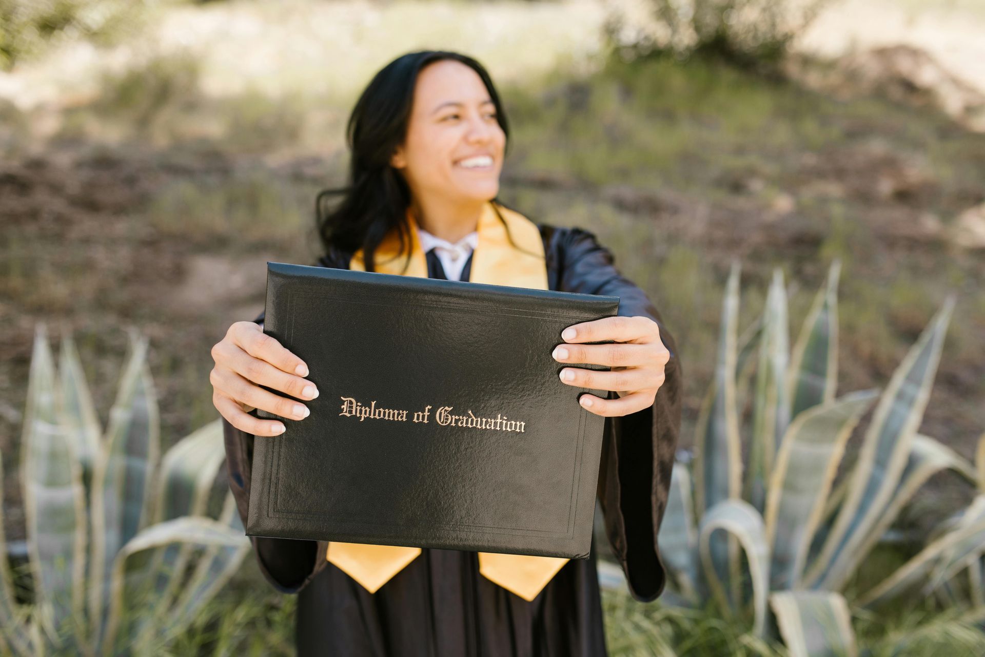 A graduate smiling and holding a black diploma cover in front of an agave plant backdrop.