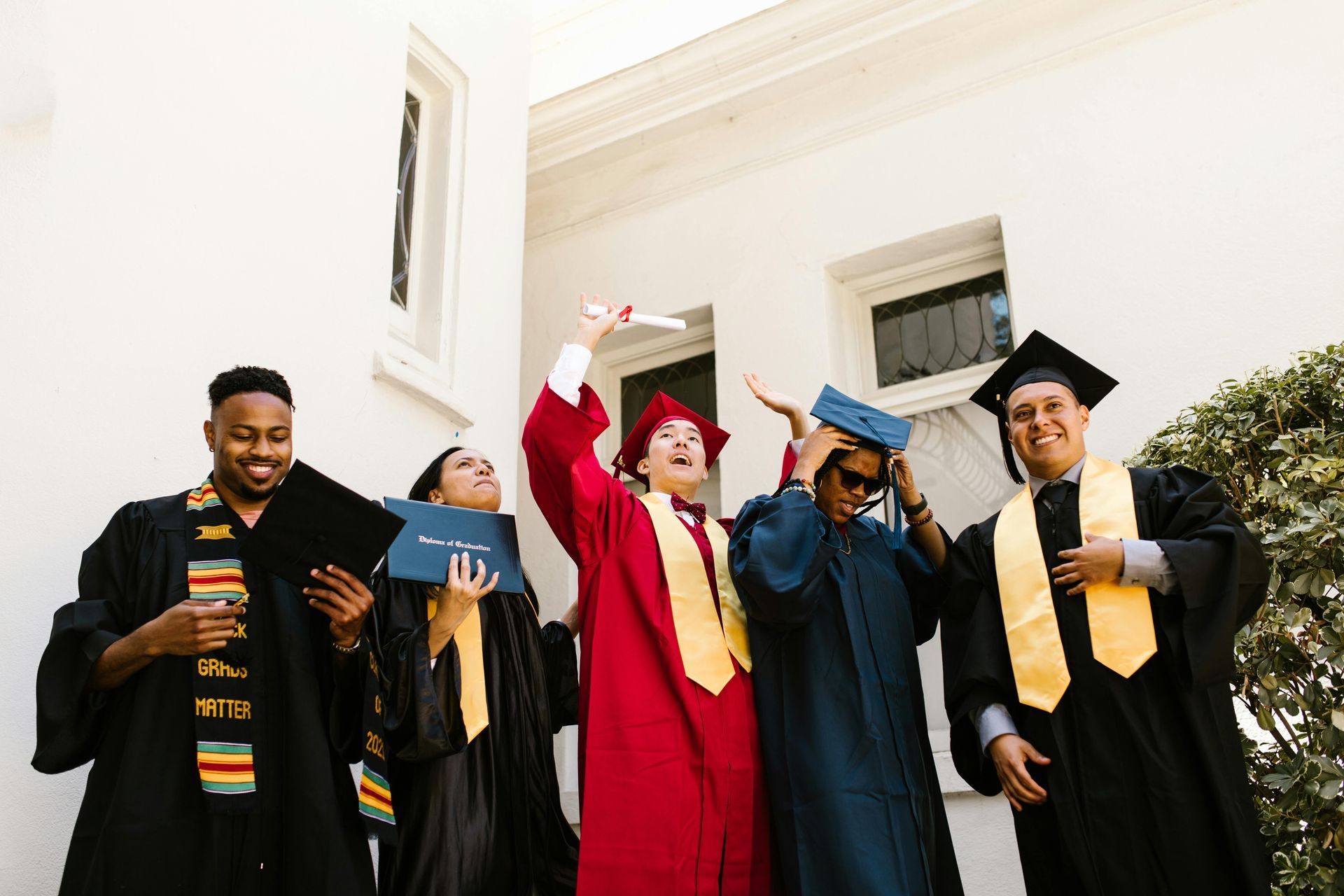 Five graduates in caps and gowns stand in front of a white building, some holding diplomas and celebrating.