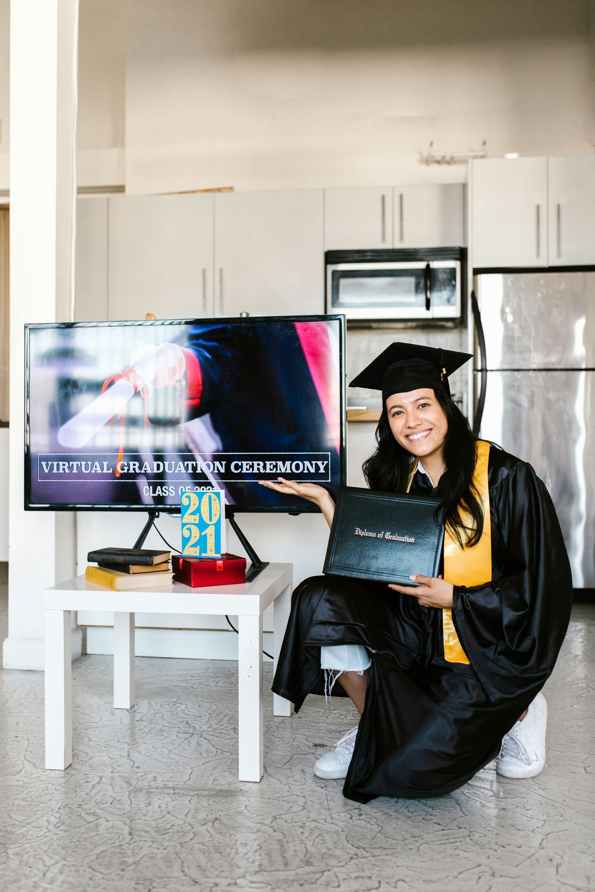 A graduate in a cap and gown holds a diploma, gesturing toward a TV displaying a 