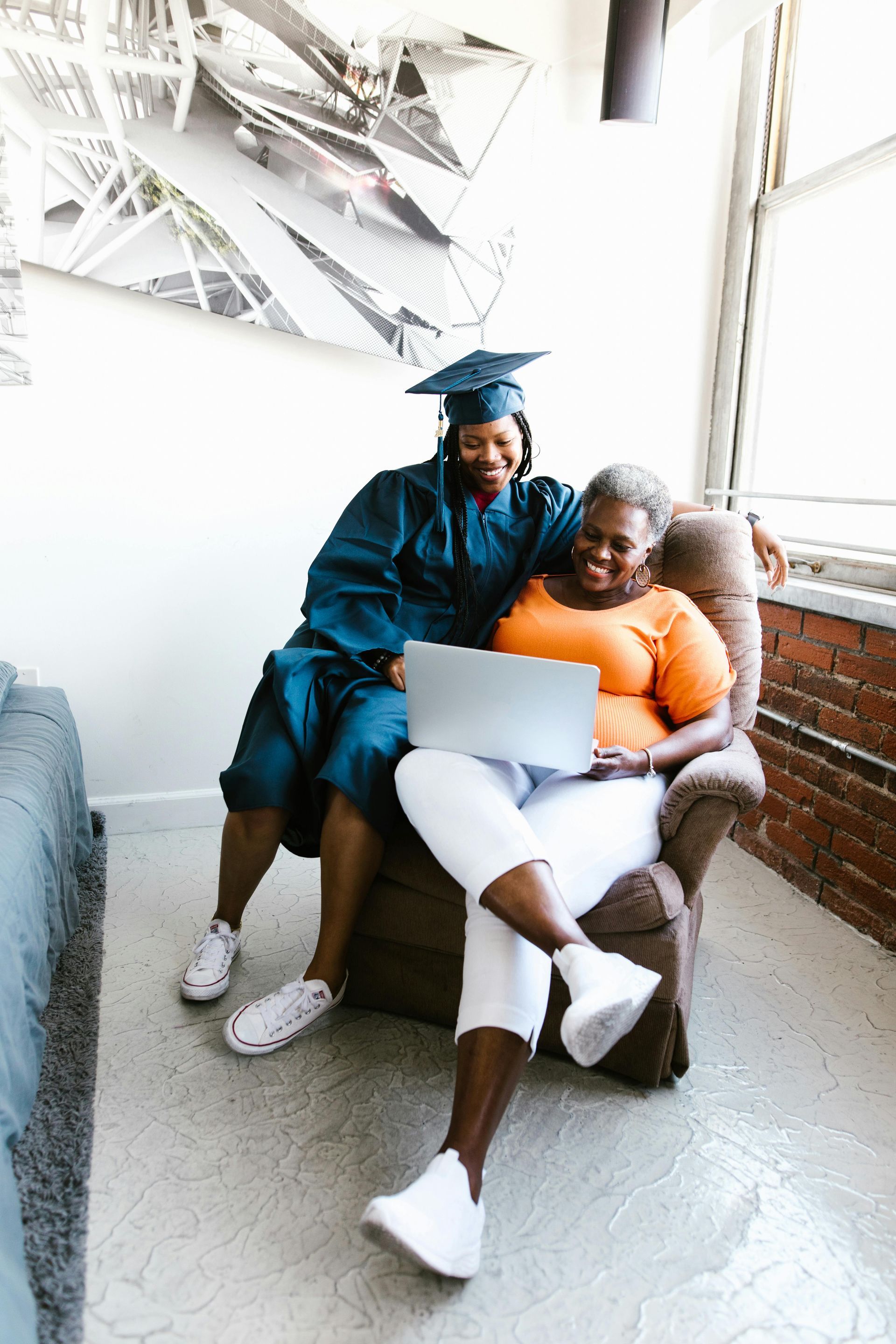 A student in graduation regalia and an adult sit in an armchair looking together at an open laptop in a sunlit room.