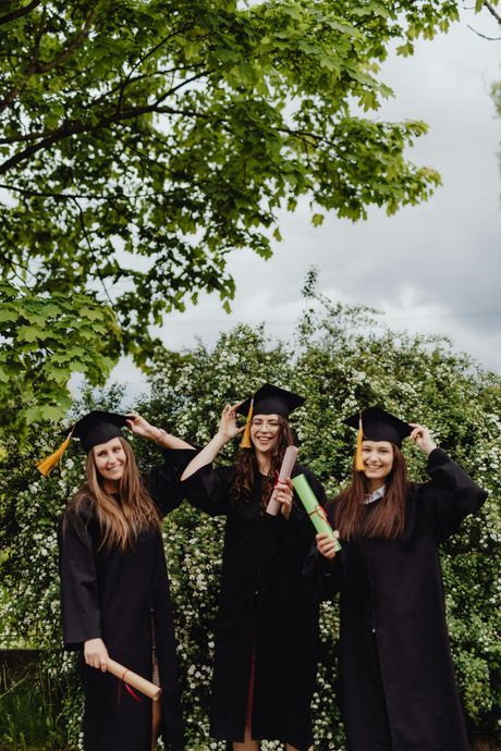 Three graduates in caps and gowns smiling and posing for a photo outdoors in front of leafy trees.