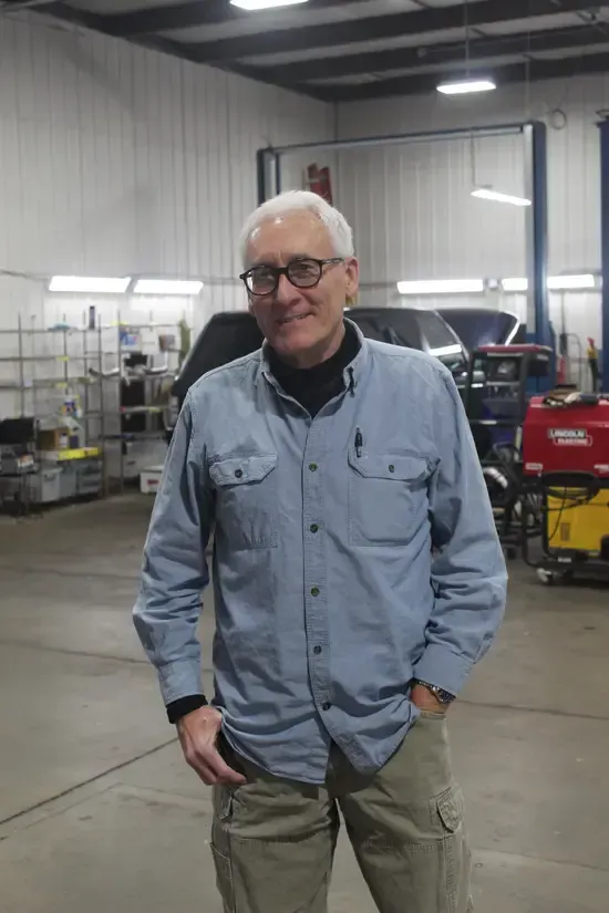 A man in a blue shirt is standing in a garage with his hands in his pockets.