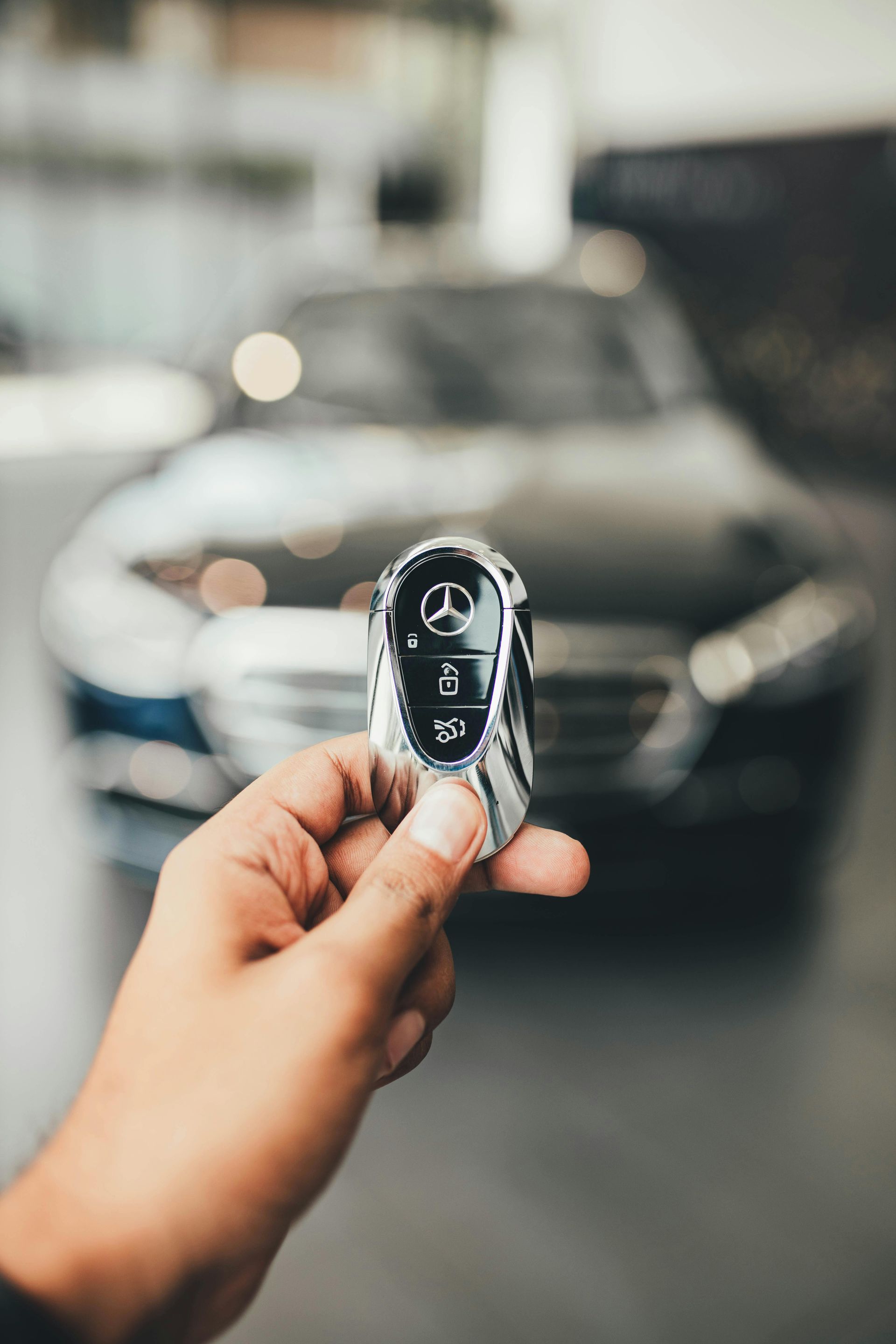Hand holding a Mercedes-Benz key fob in front of a blurred black car.