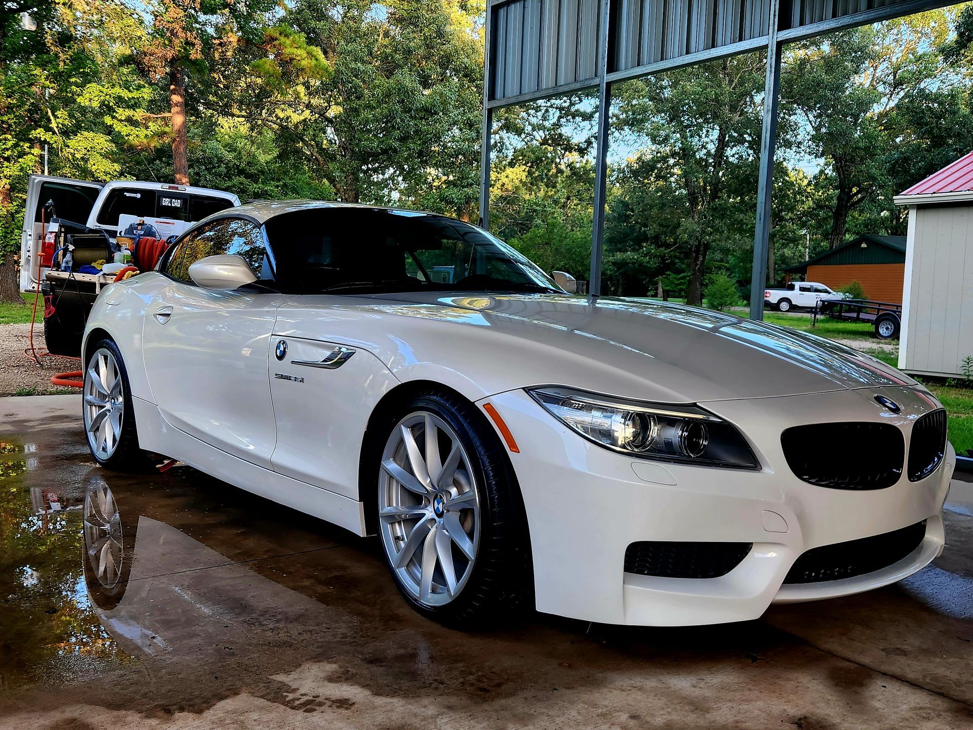 White BMW convertible parked under a carport with a wet driveway and a red truck in the background
