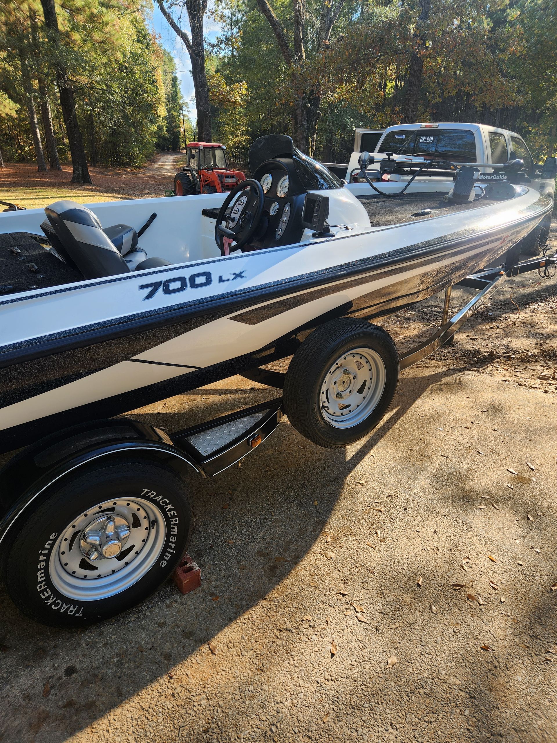 Bass boat on a trailer; black, white, and silver. Outdoors in sunlight, beside trees and a van.