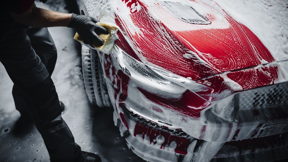 Person washing a red sports car with a sponge and soapy water.