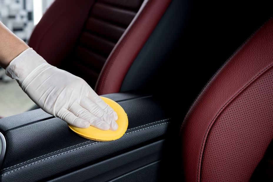 Person in a white glove cleaning black car armrest with a yellow sponge, against a red leather seat.