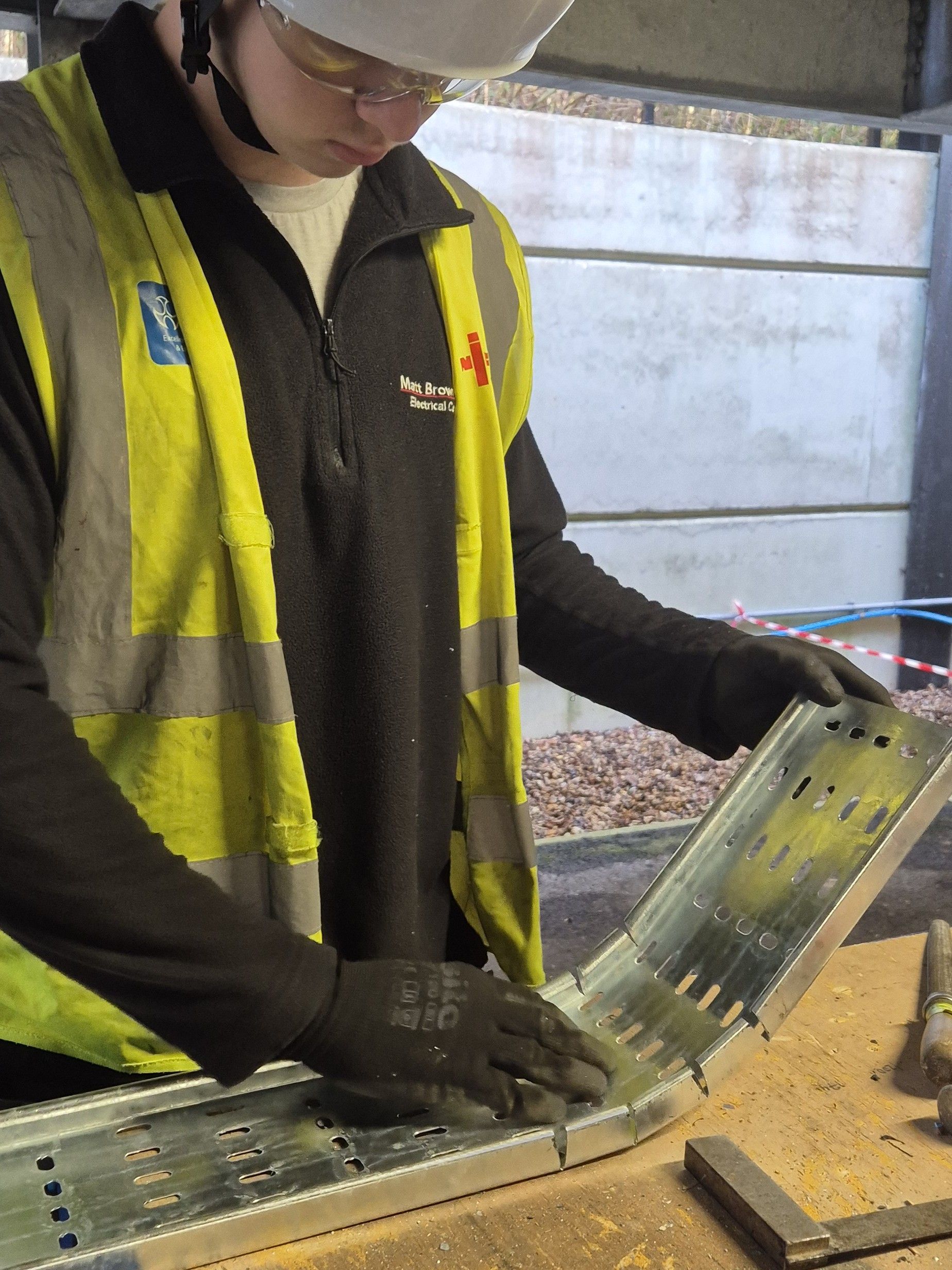 Electrical Apprentice with PPE fabricating galvanised steel cable tray bend for a commercial electrical installation at Matt Browning Electrical. NICEIC Approved Contractor and ECA Electrical Contractors Association Logos