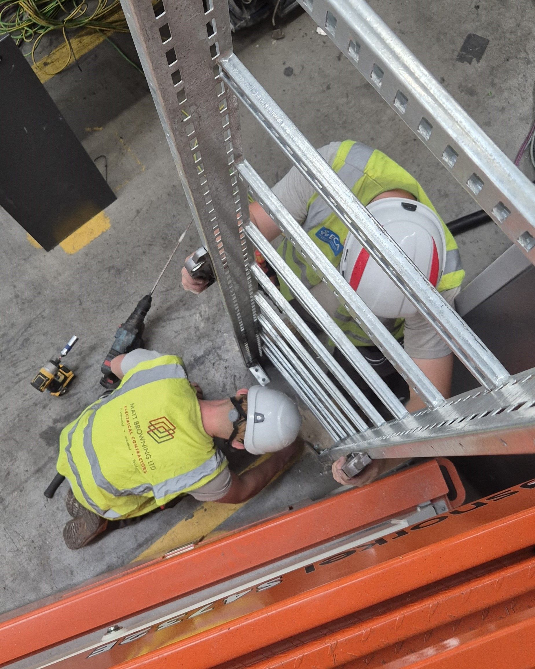 An aerial view of an electrician and electrical apprentice installing a vertical cable ladder section to deliver electrical power to a new machine installation. The machine can be seen in the background. They are wearing PPE & hi-vis while working in a commercial industrial building as part of a factory electrical fit-out by the skilled electrical team at Matt Browning Electrical