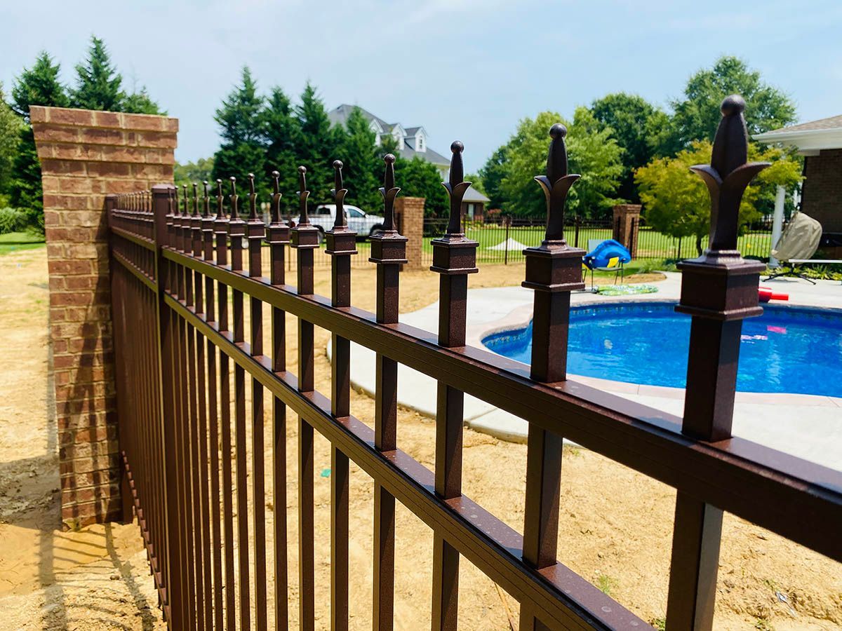 Brown metal fence beside a blue swimming pool in a sunny backyard with trees and brick pillars