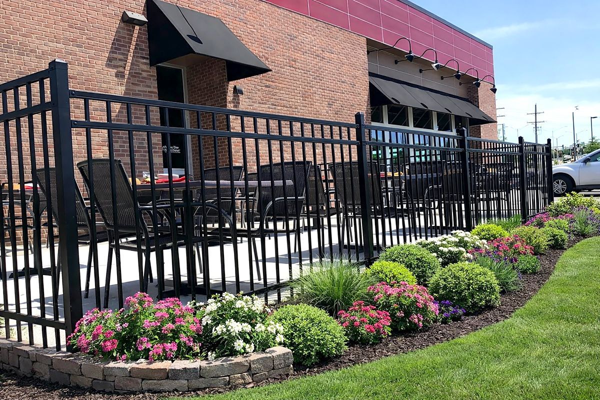 Brick restaurant patio with black fence, red awning, and pink flower beds along a sidewalk