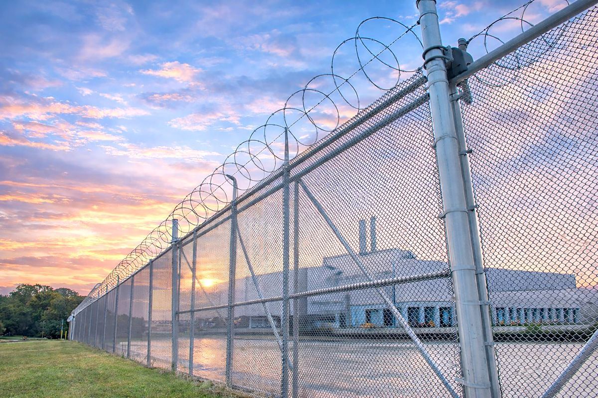 Chain-link prison fence topped with razor wire at sunset, with a low building in the background.