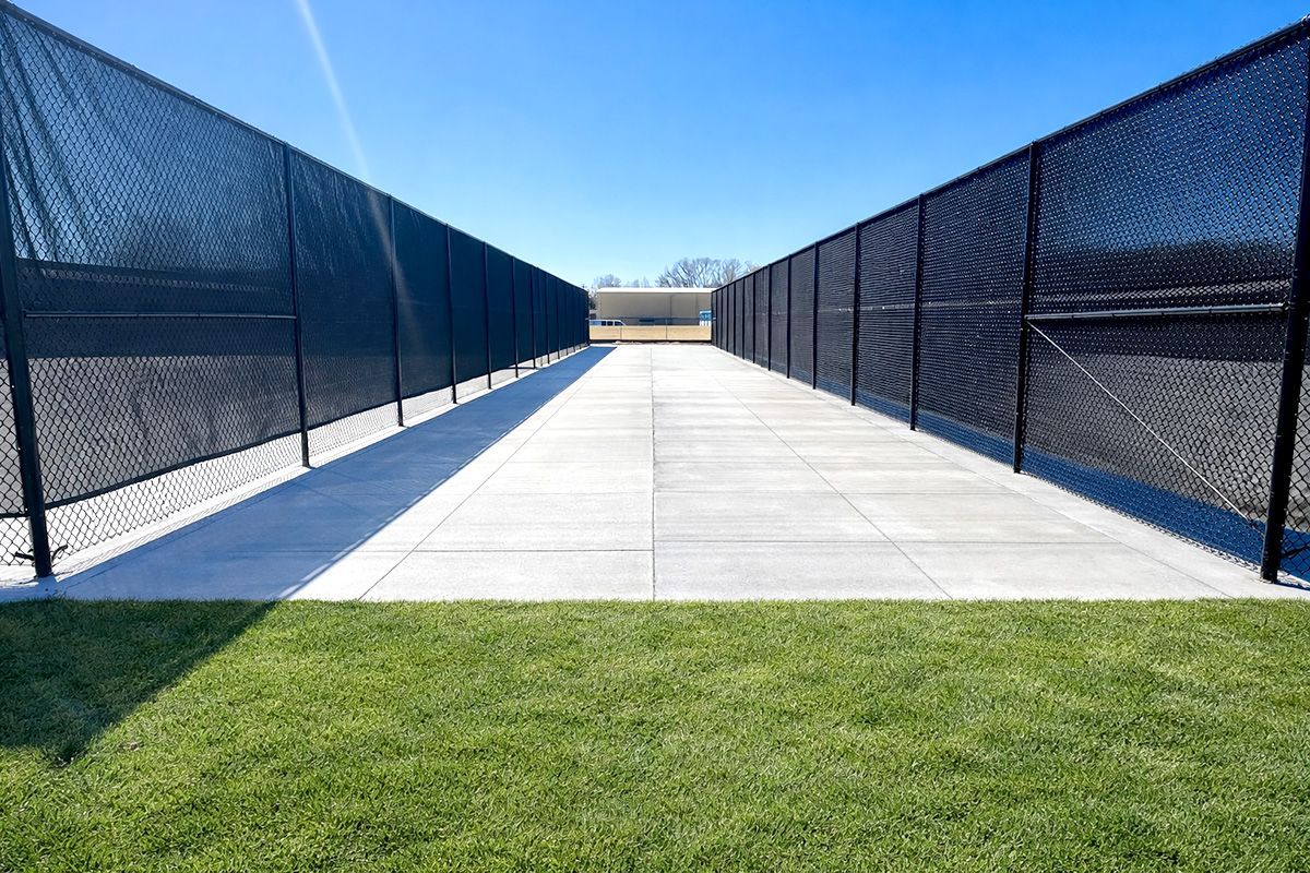 Empty concrete walkway between tall black chain-link fences with green grass in the foreground