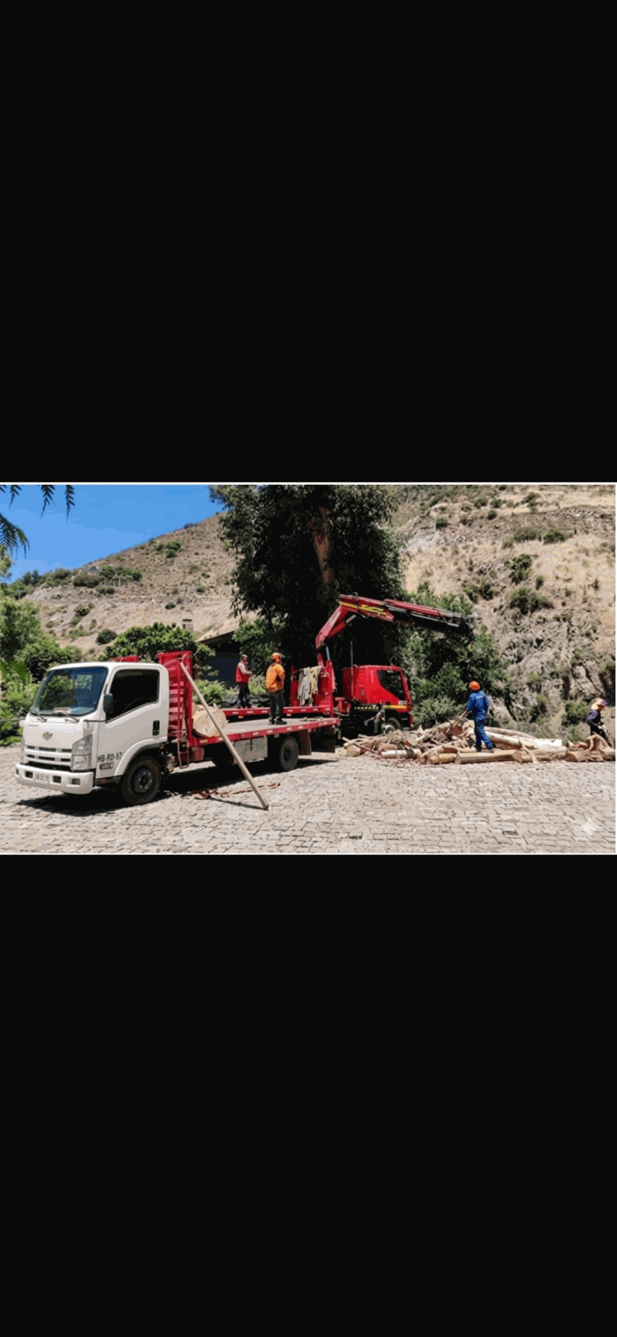 Una excavadora y un camión blanco junto a una carretera rocosa, con trabajadores bajo un cielo azul despejado.