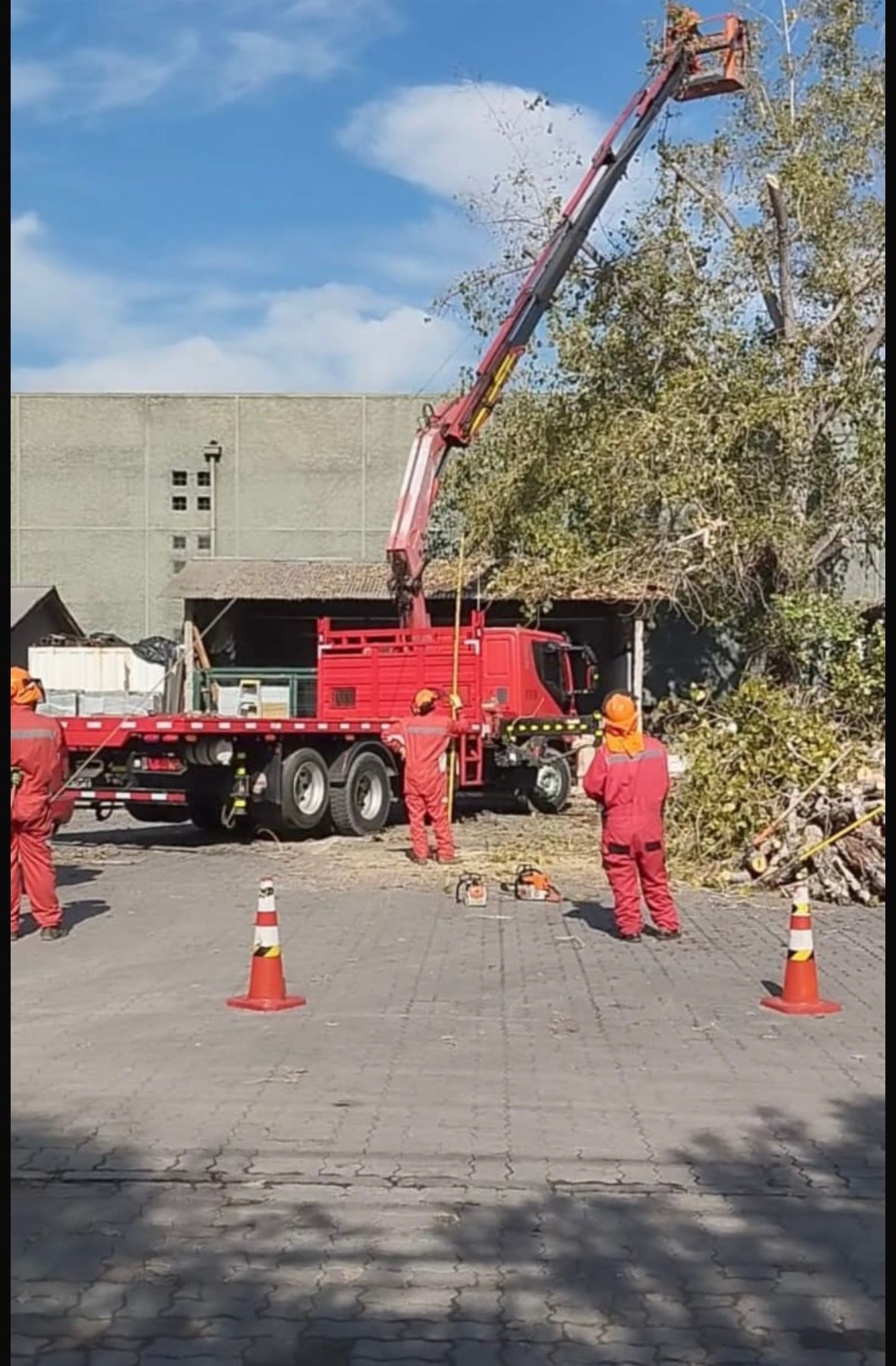 Grúa camión roja levantando un brazo largo junto a un edificio, con trabajadores y conos de tráfico cerca